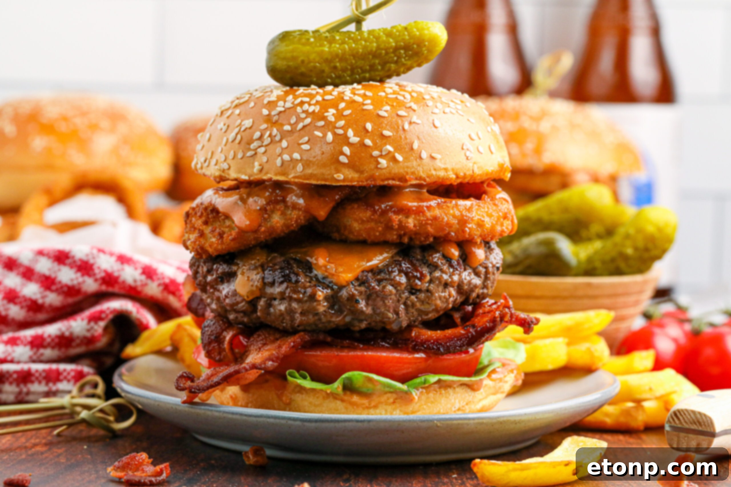 side view of the Cowboy Burger on a white plate that is fully loaded with toppings with a small bowl of pickles and fries in the background
