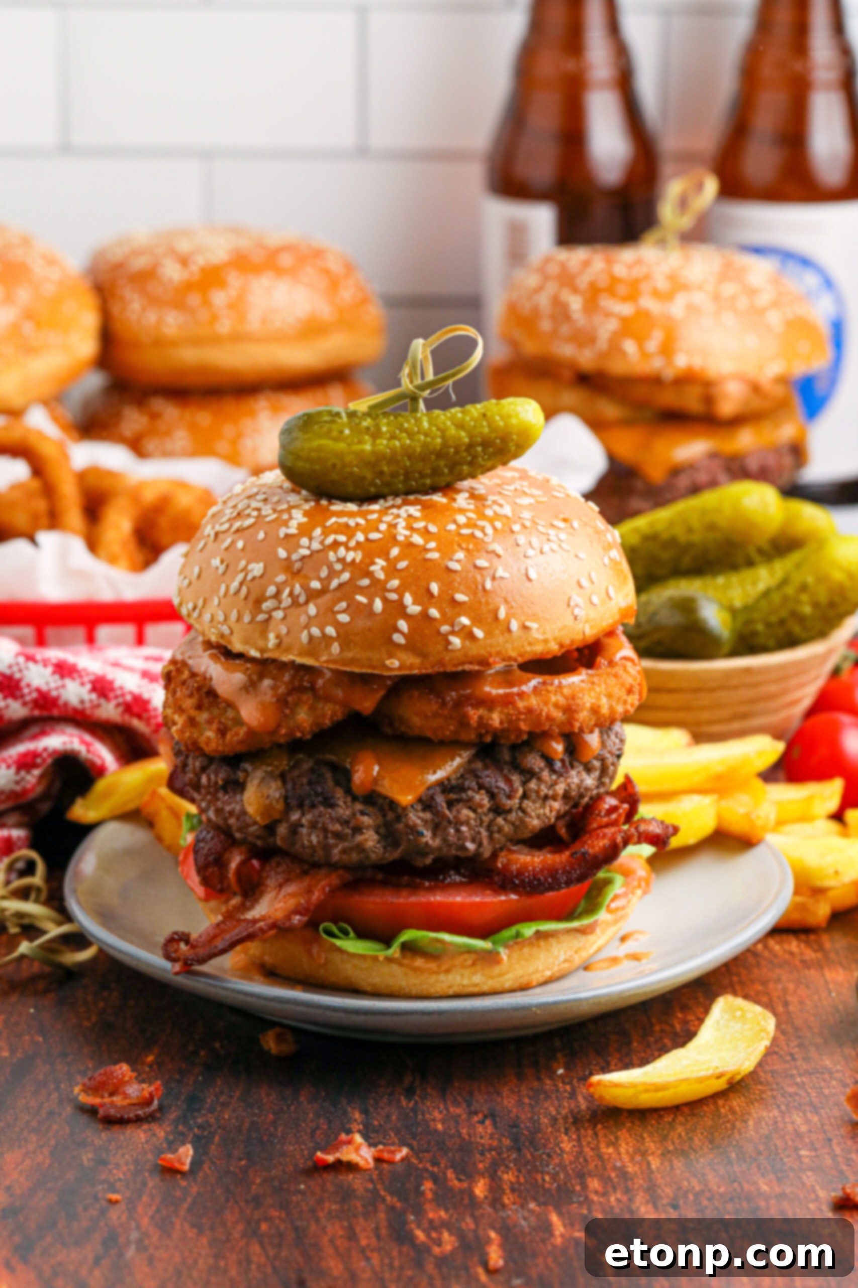 far away shot of the Cowboy Burger on a white plate that is fully loaded with toppings with a small bowl of pickles and fries in the background