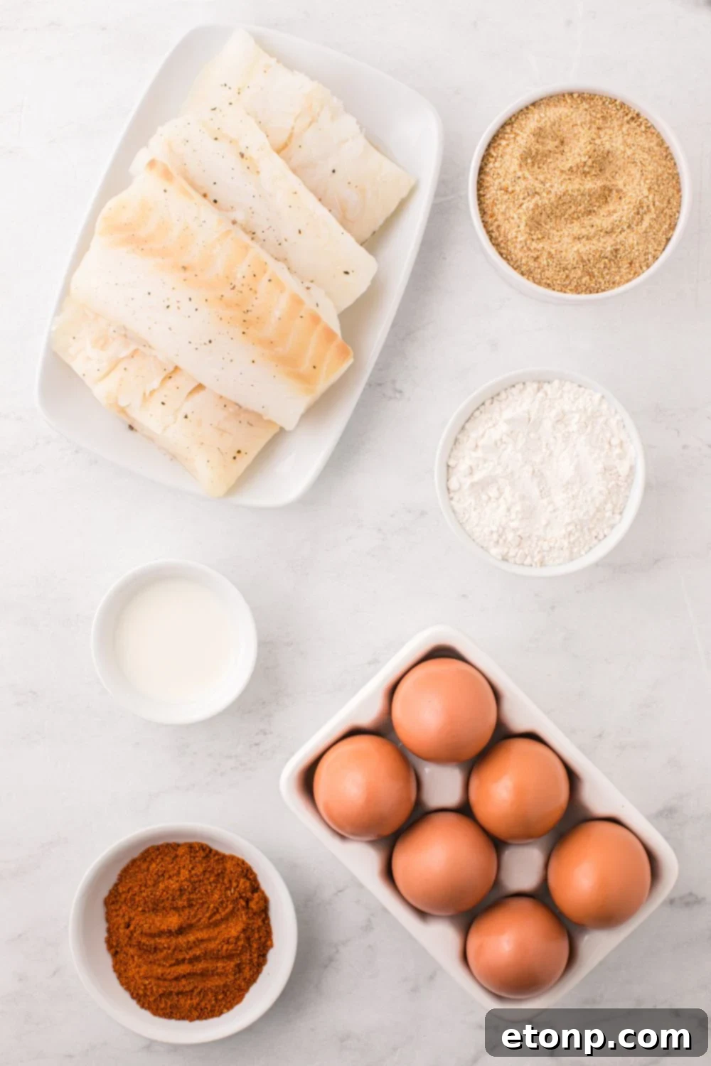 Overhead view of fresh ingredients laid out on a counter, including white fish fillets, flour, eggs, buttermilk, breadcrumbs, and Old Bay seasoning, symbolizing the start of the cooking process.