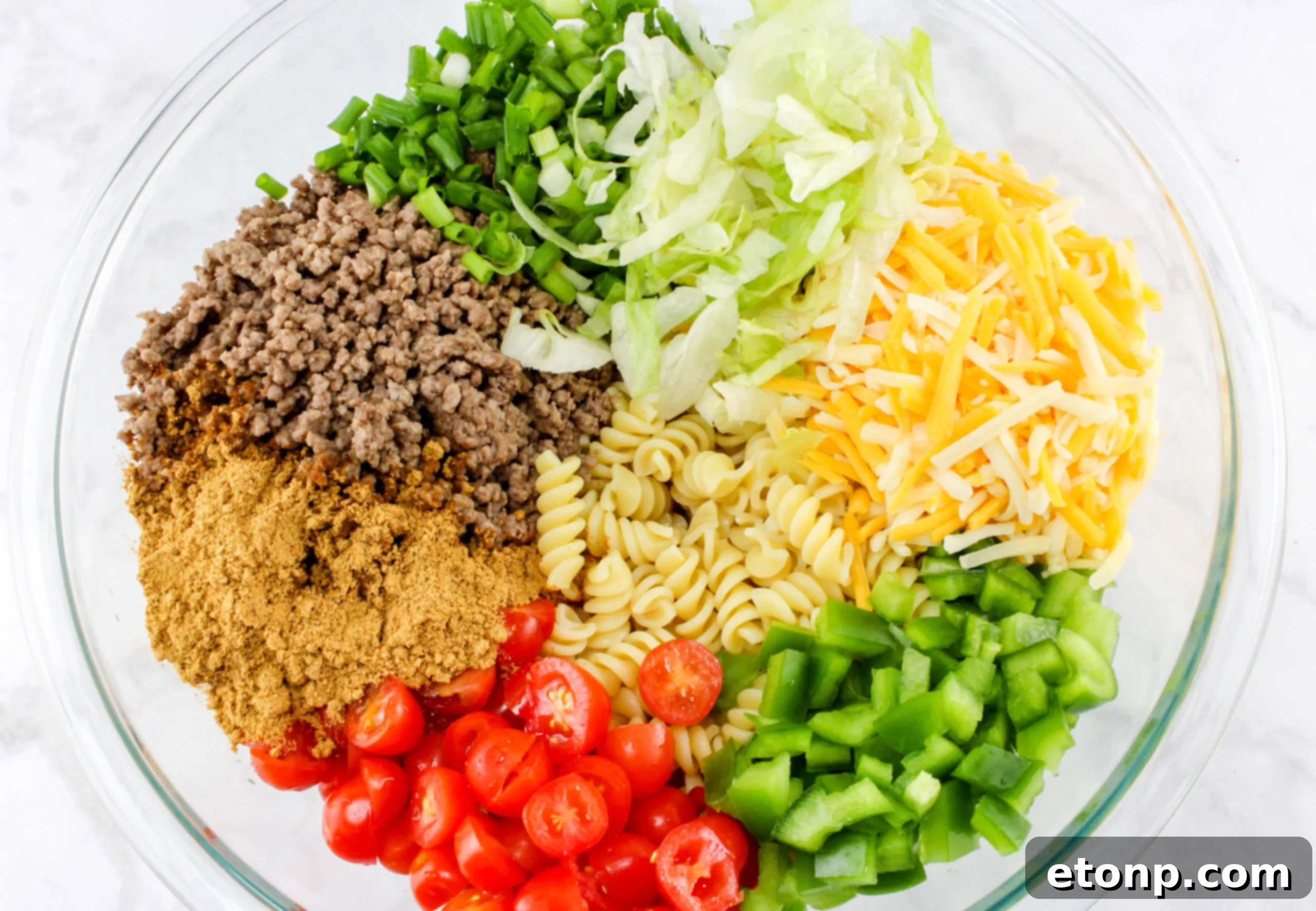An overhead shot showcasing the colorful ingredients of the pasta salad, including spiral pasta, ground meat, fresh lettuce, and cherry tomatoes, all mixed and ready.