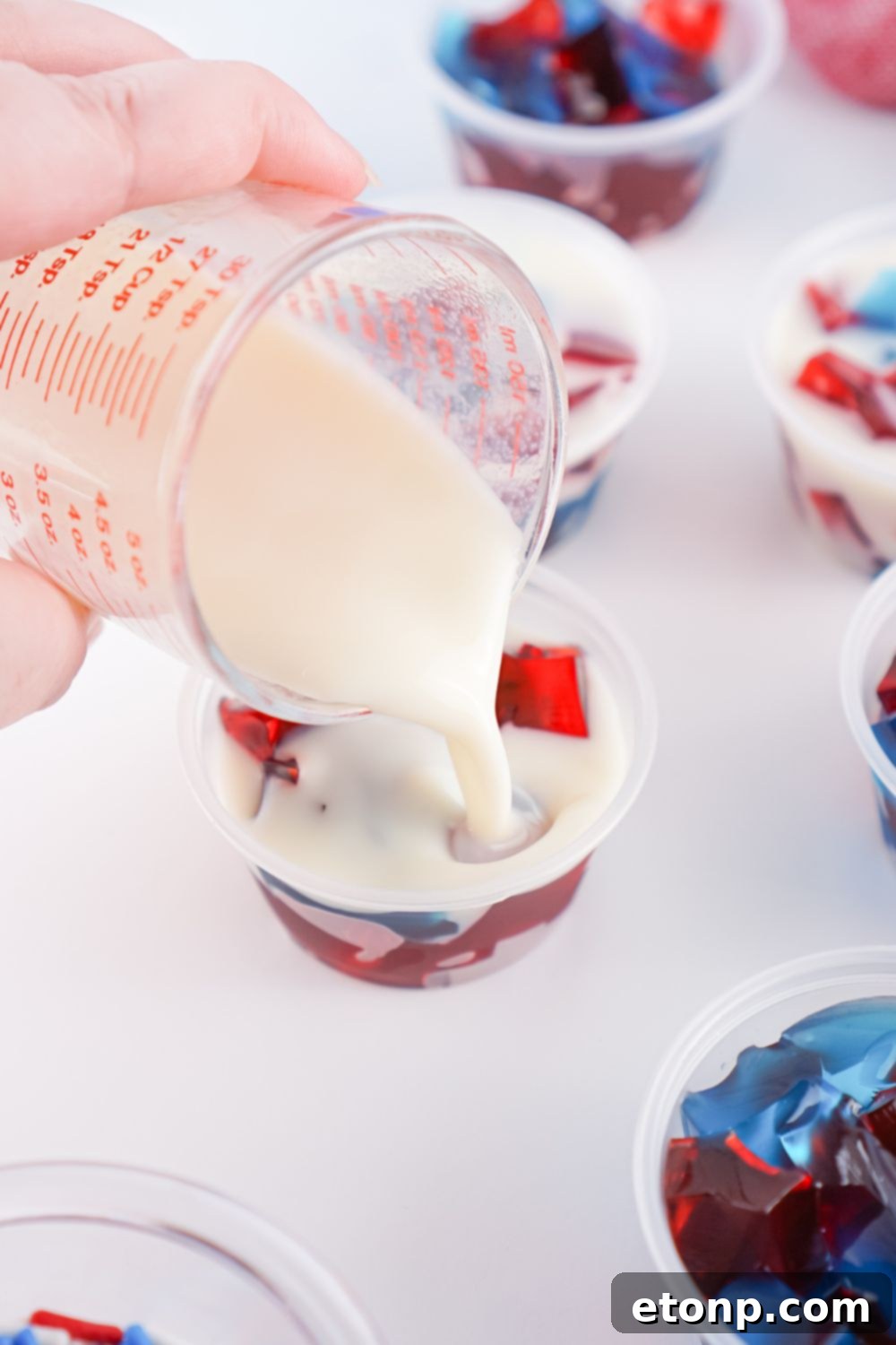 The creamy white jello mixture being poured over the red and blue jello cubes in a shot container, creating the distinctive stained glass layers.