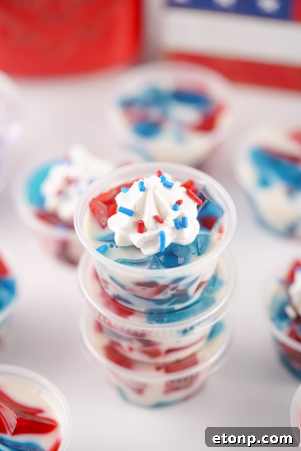 Red, White, and Blue Jello Shots stacked, with an American flag visible in the blurred background, emphasizing their patriotic theme and portability.