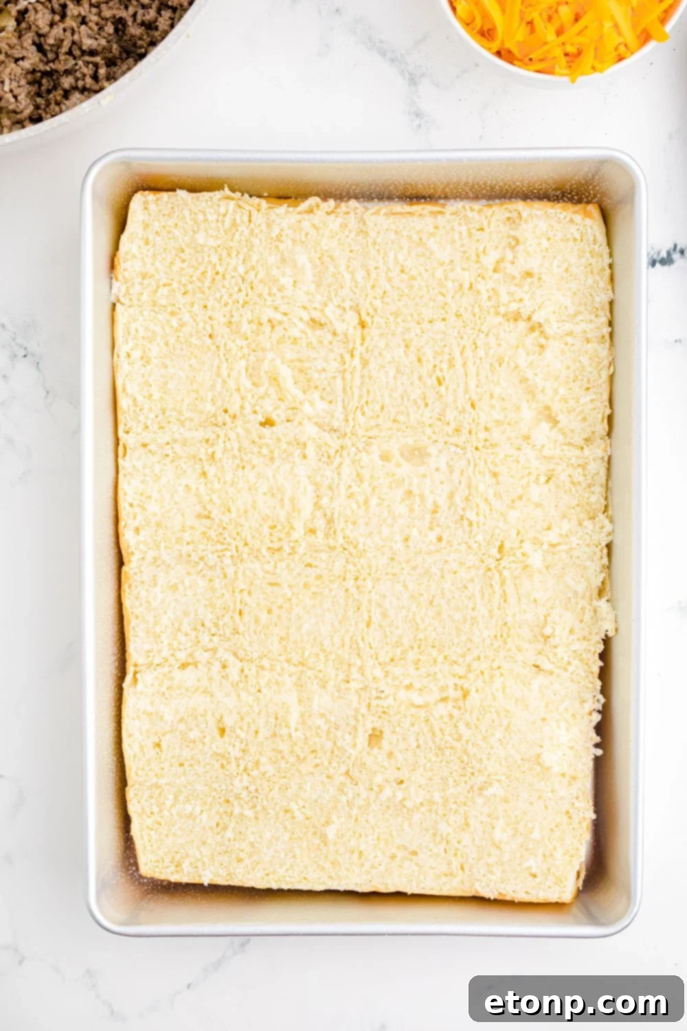 A pristine baking pan holds a sheet of Hawaiian rolls, expertly sliced in half horizontally, forming the base for the delightful Classic Cheeseburger Sliders.
