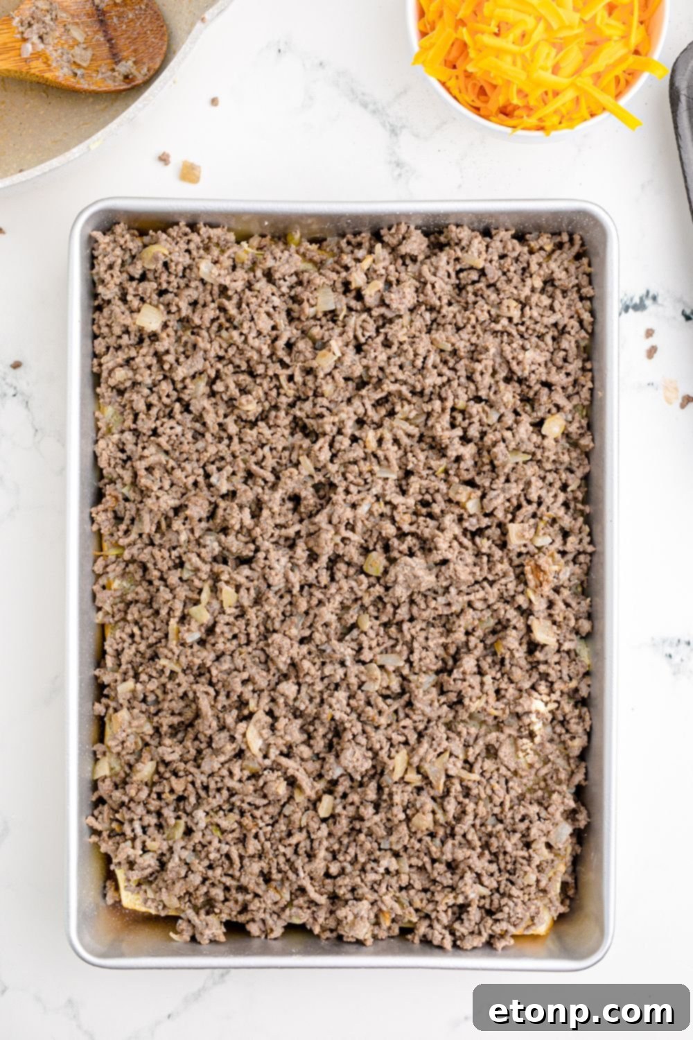 A generous layer of seasoned ground beef spread evenly over the bottom halves of slider buns, nestled together in a baking tray, ready for the cheese and top buns.