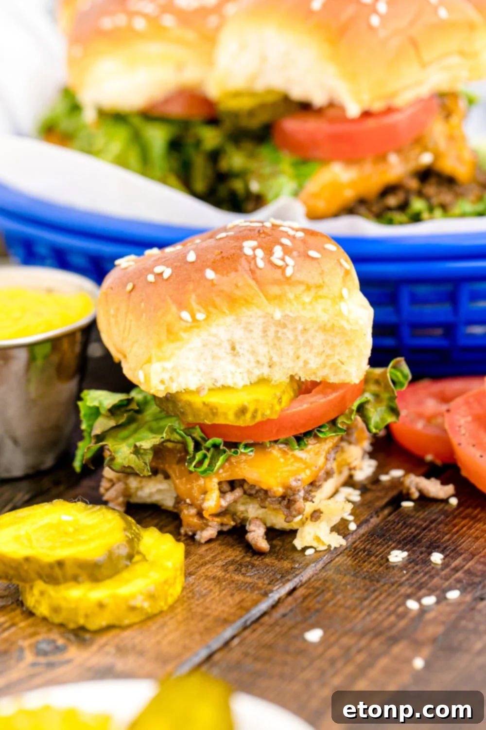 A single Classic Cheeseburger Slider elegantly placed on a light-colored table, accompanied by a small array of fresh sliced pickles and tomatoes, ready to be enjoyed.