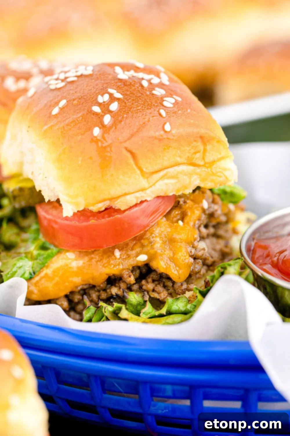 A single Classic Cheeseburger Slider elegantly presented in a small, blue-checkered basket, with a tiny bowl of bright red ketchup positioned invitingly to the side, highlighting its snackable perfection.