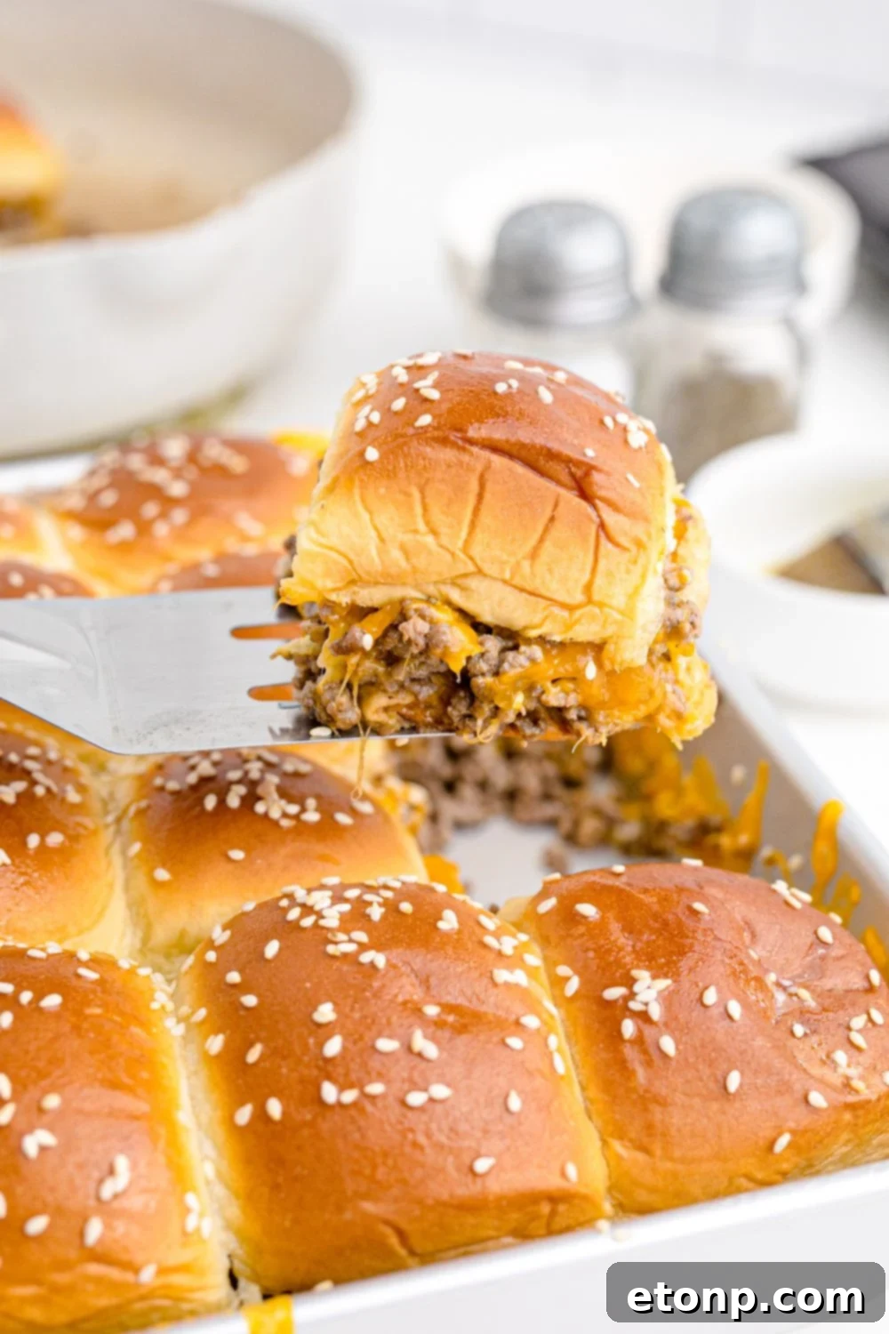 A hand gently lifting a Classic Cheeseburger Slider from a baking pan filled with many more, showcasing the melted cheese and soft buns, ready for serving.