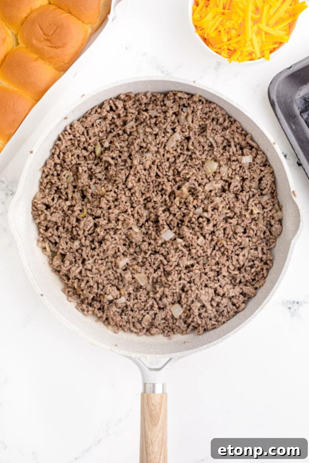 A kitchen counter arrangement featuring a skillet of cooked ground beef, a stack of soft slider rolls, and a bowl of shredded cheese, all the key components for assembling Classic Cheeseburger Sliders.