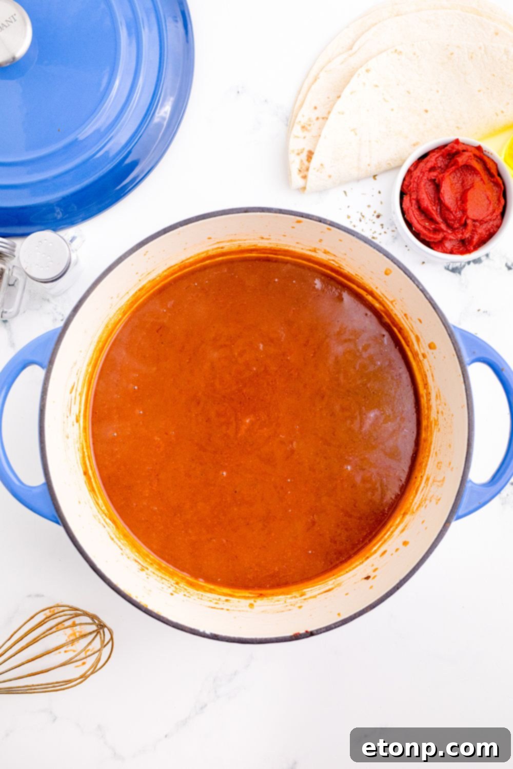 From Scratch Enchilada Perfection 9 Homemade Enchilada Sauce in a pot sitting on table with whisk to the side of pan, simmering to ideal thickness