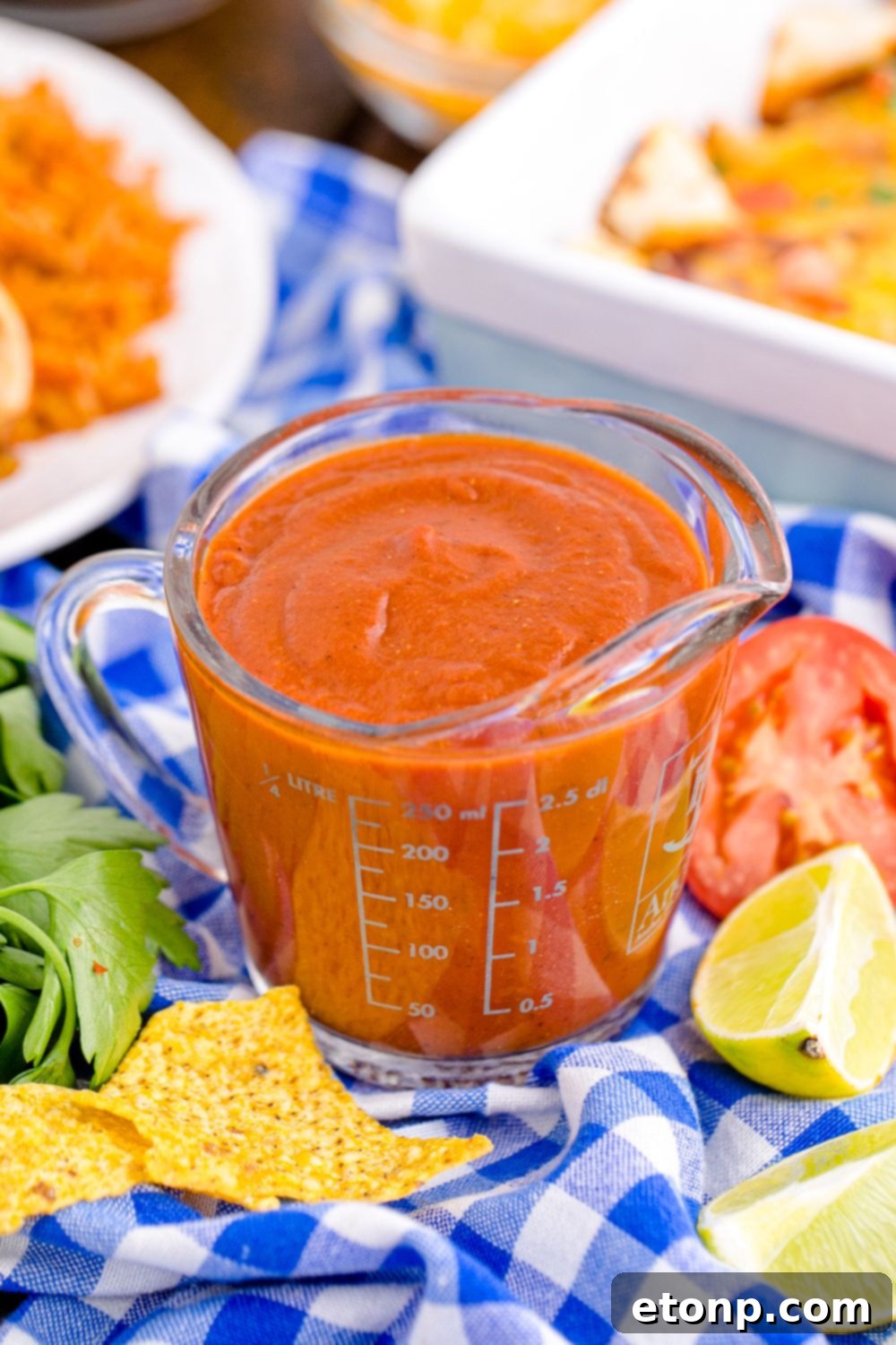 From Scratch Enchilada Perfection 10 measuring cup with enchilada sauce sitting on table with tomatoes, cilantro, chips and more scattered around on table, perfect for a gathering