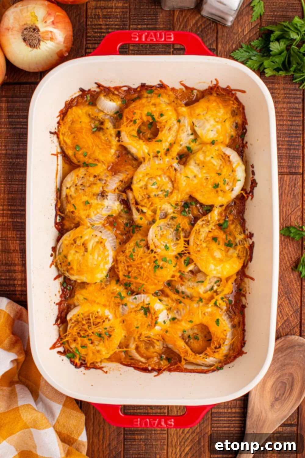 Cheesy Tennessee Onion Bake 5 Overhead shot of Cheesy Tennessee Onions in a baking dish on a wooden table, surrounded by fresh herbs and raw onions.