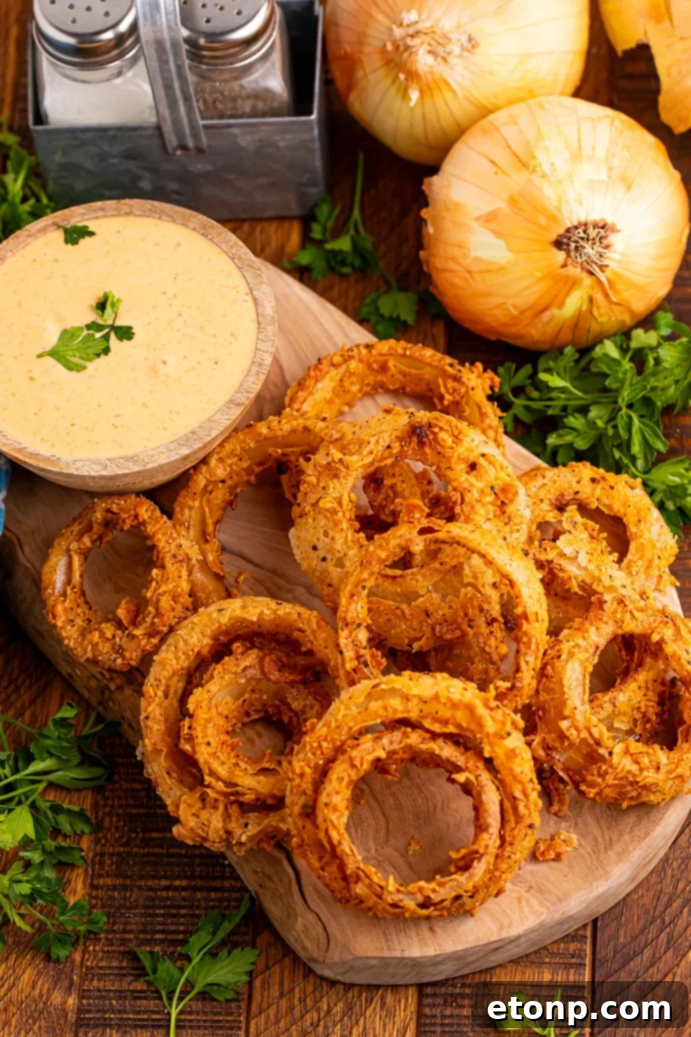 Golden Crispy Homemade Onion Rings 21 looking down at table to see a wooden tray with parsley, whole onions, and a bowl of sauce topped with parsley