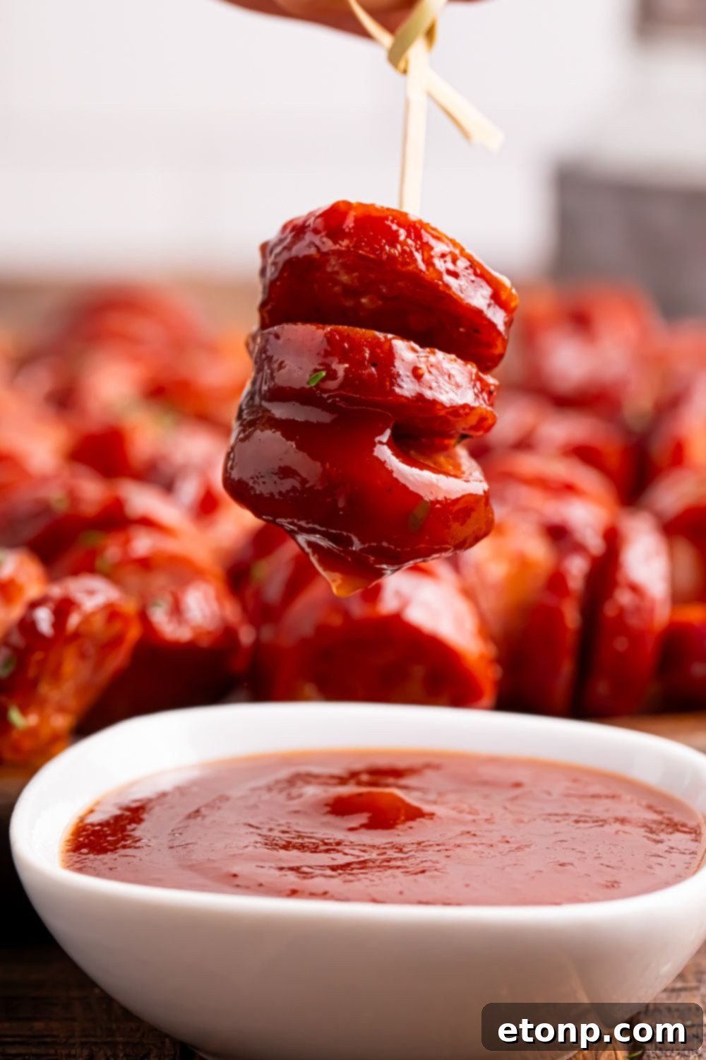 BBQ Hasselback Smoked Sausages dipped in sauce being held above the bowl 