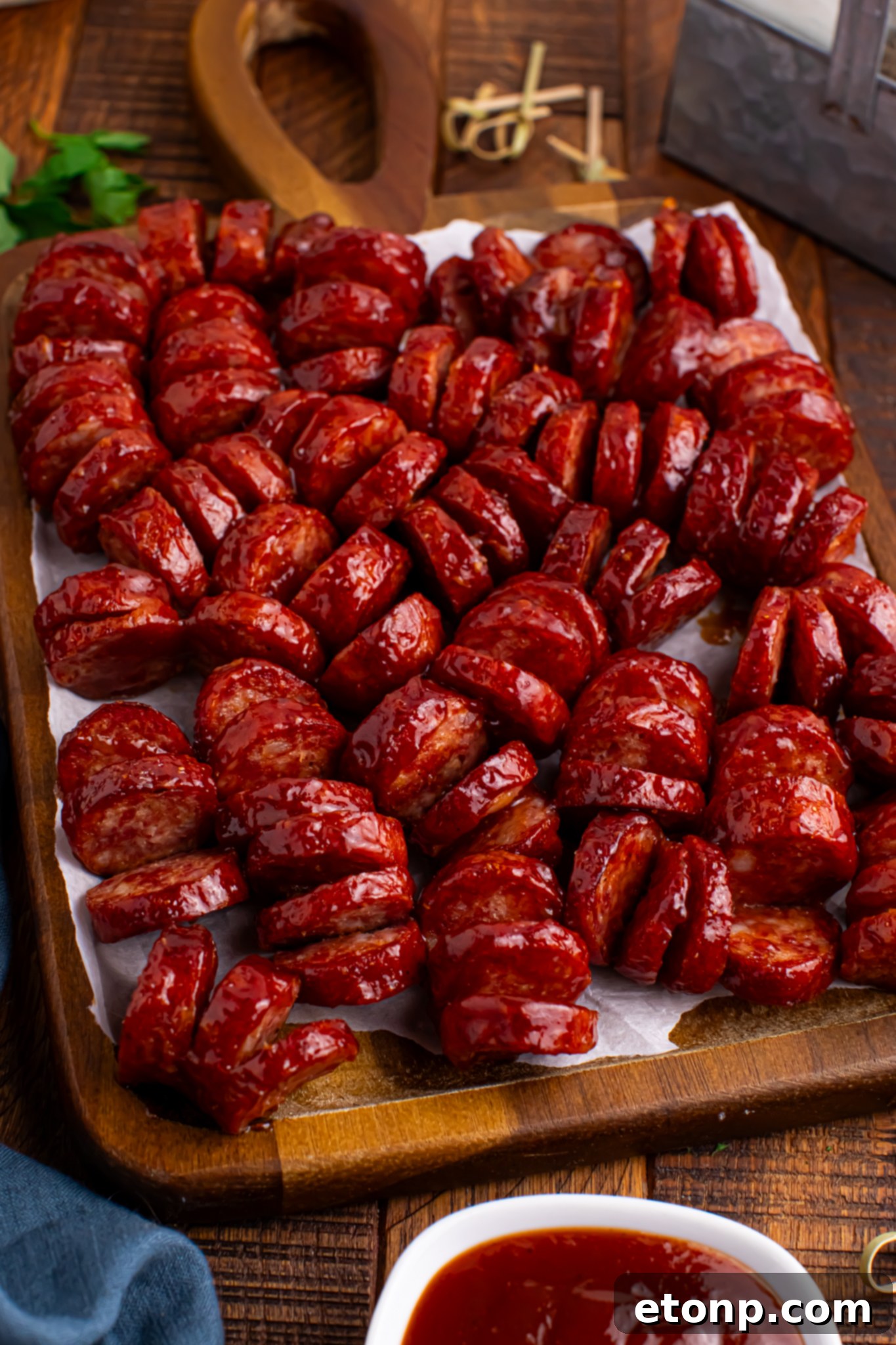 close up view of BBQ Hasselback Smoked Sausages spread out on a dark wood cutting board with a white bowl of bbq in front 