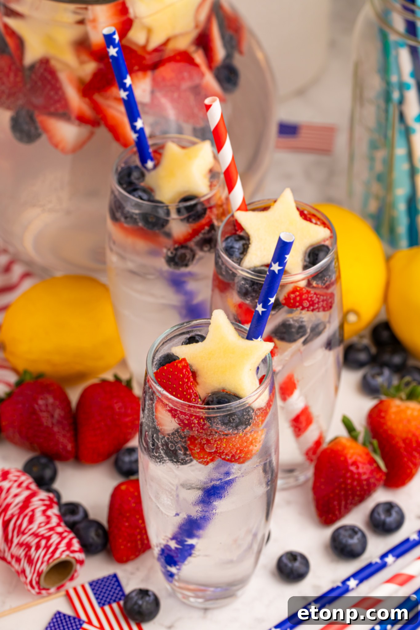 Liberty Lemon Sparkle 2 3 tall clear glasses of Patriotic Sparkling Lemonade with flag straws and with fruit scattered on table