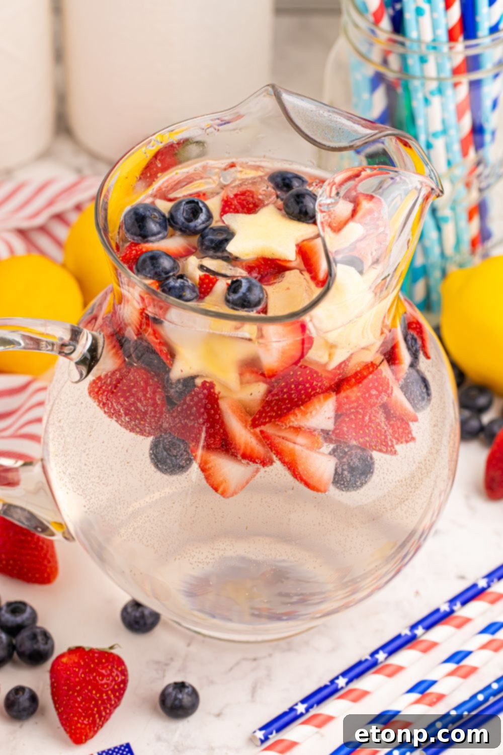 Liberty Lemon Sparkle 18 Patriotic Sparkling Lemonade in a container on table with blueberries and strawberries, with paper patriotic straws scattered on table
