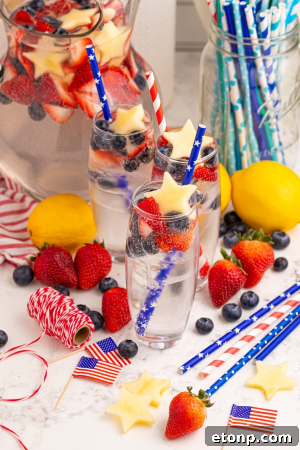 Liberty Lemon Sparkle 21 Patriotic Sparkling Lemonade in glasses with pitcher of lemonade to side. Fresh fruit scattered around on table with little American flags, and ribbon