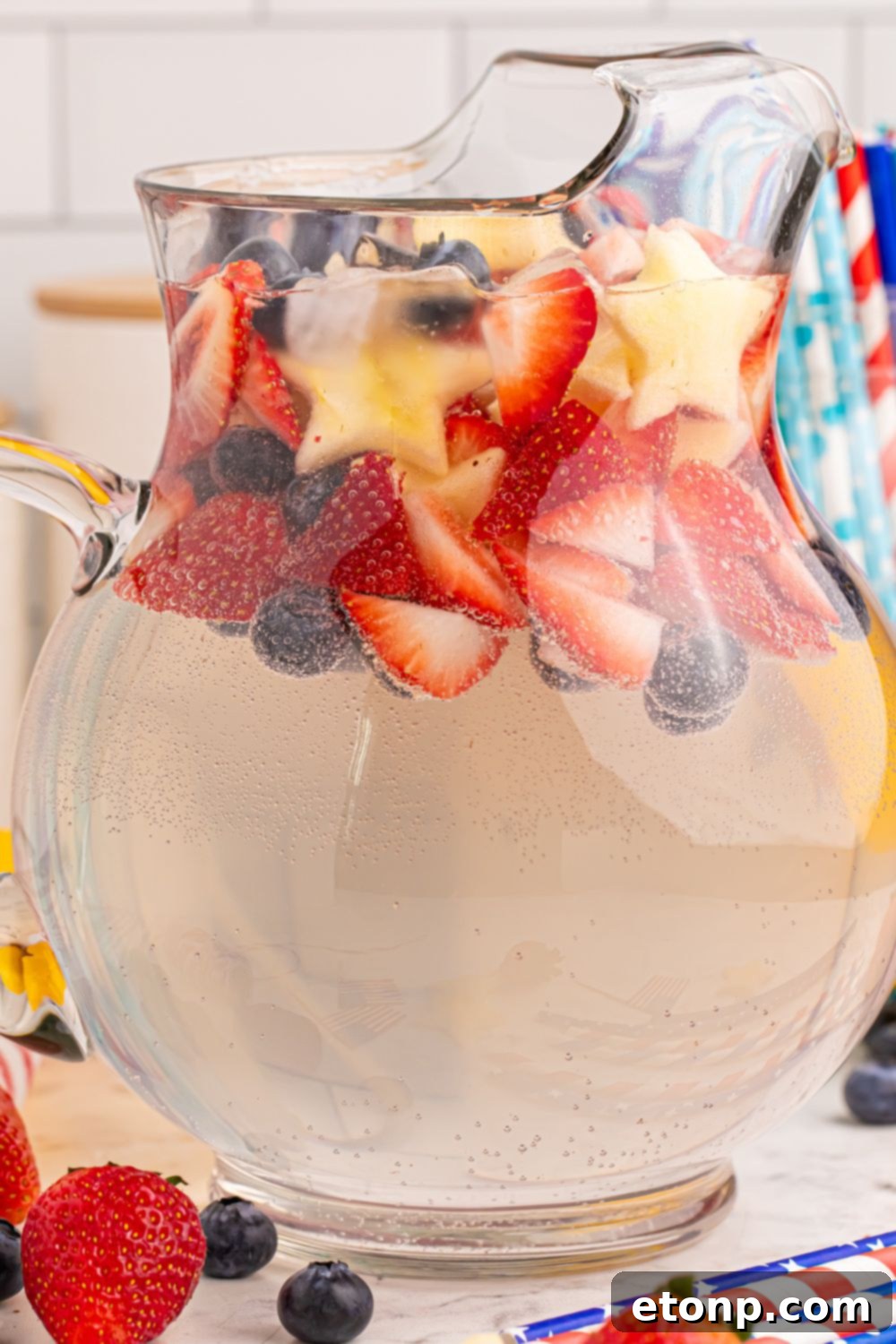 Liberty Lemon Sparkle 6 side view of glass pitcher with strawberries, blueberries fruit around the table for the Patriotic Sparkling Lemonade