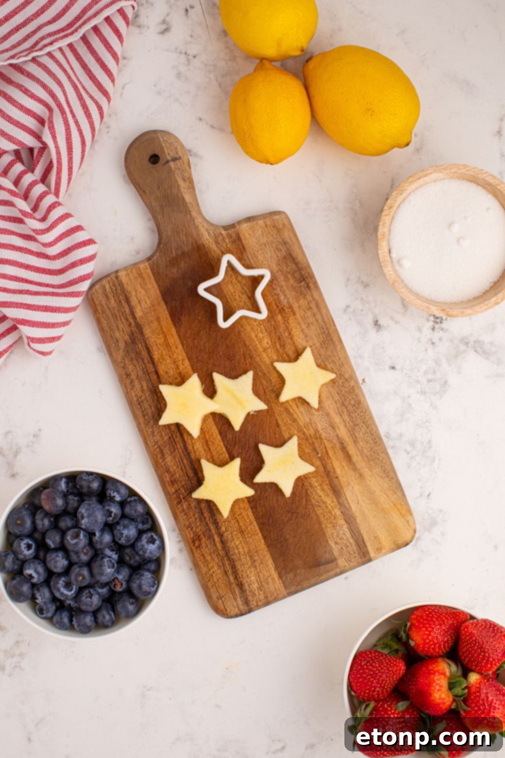 Liberty Lemon Sparkle 9 cutting board with apples cut to the shape of stars, and a bowl of sugar, blueberries ,and strawberries to the side