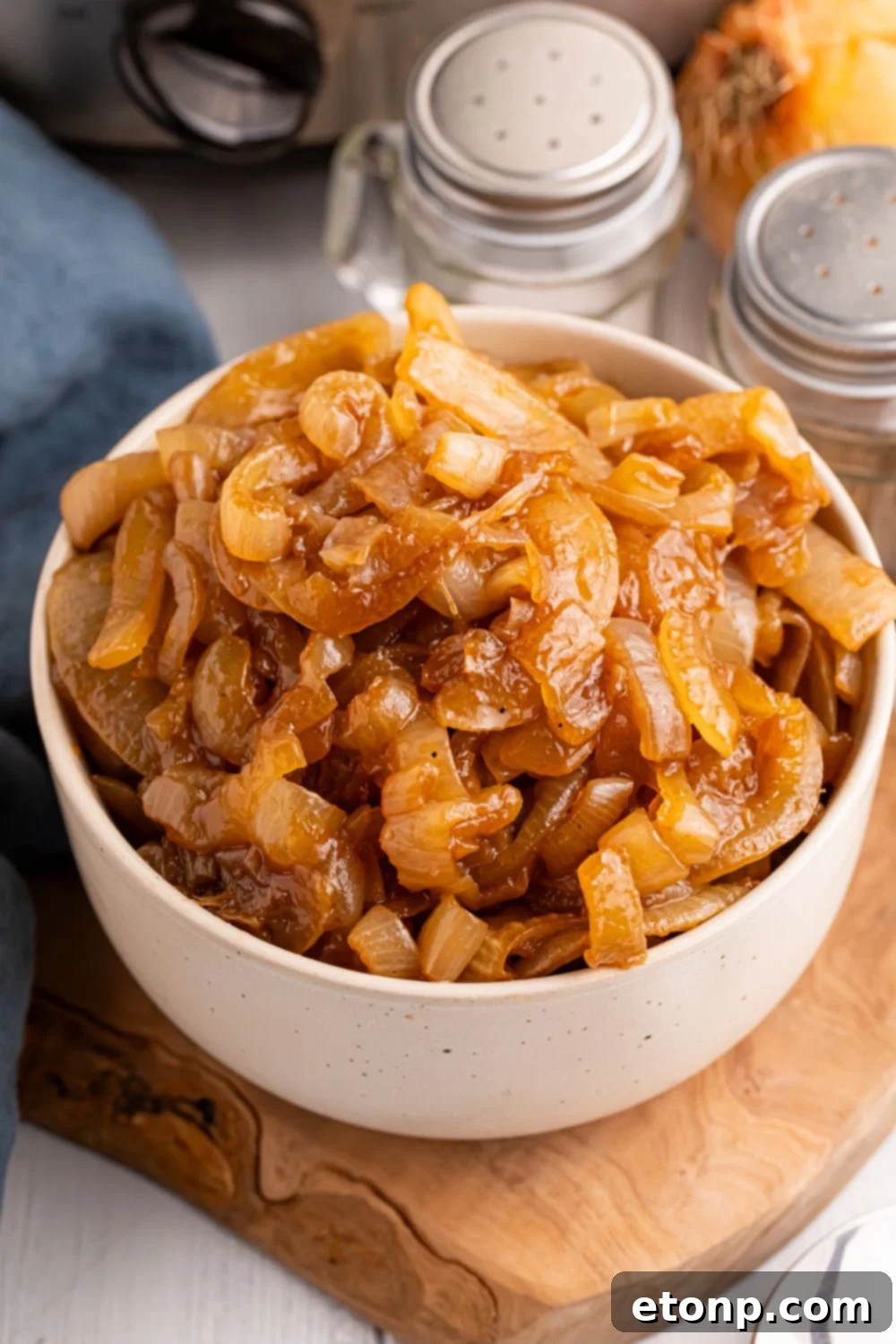 CrockPot Caramelized Onions in a bowl on a cutting board on the table. Salt, pepper, and onion to the side of the container 