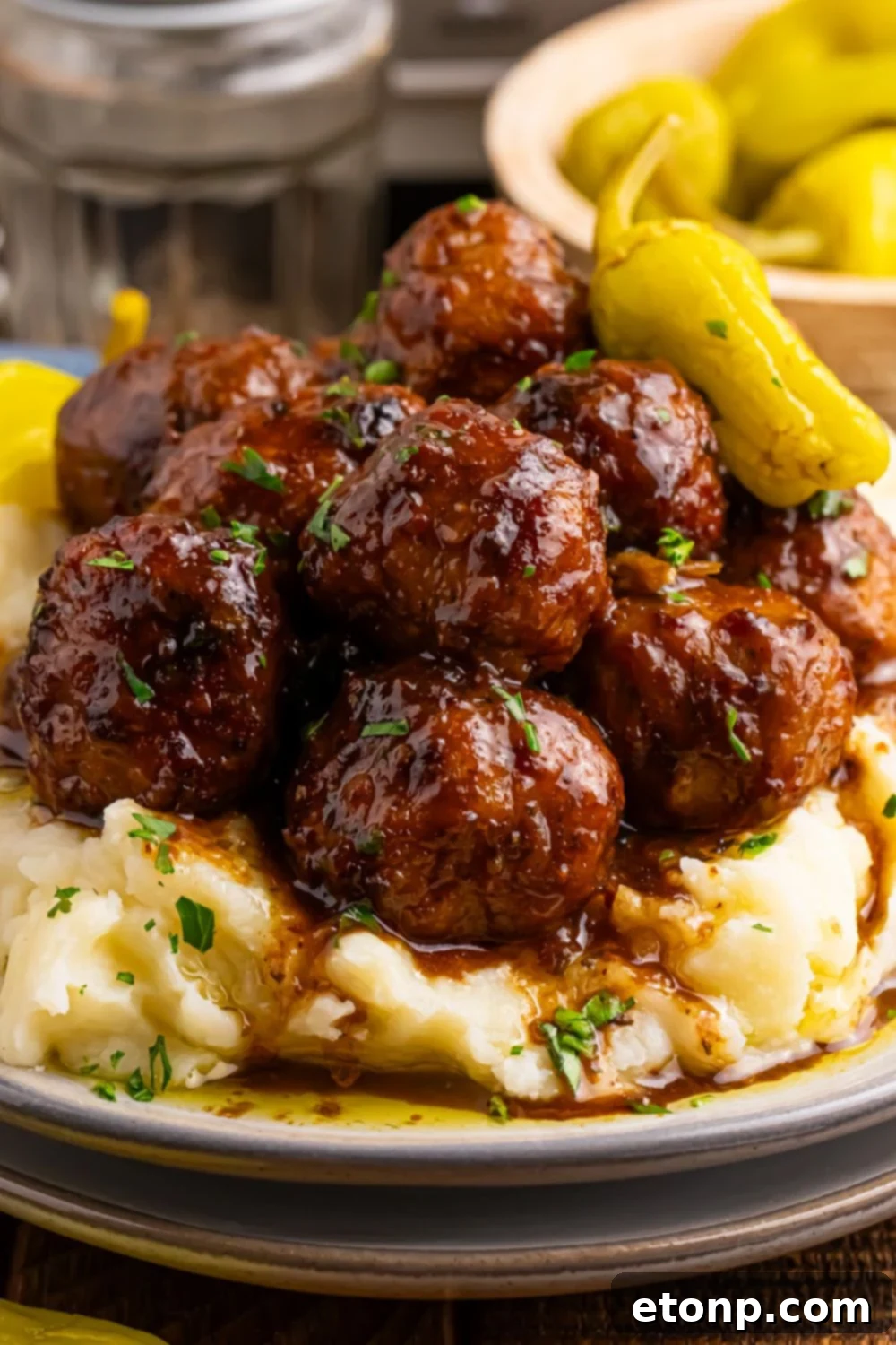 5 Ingredient Crockpot Mississippi Meatballs 20 Crockpot Mississippi Meatballs piled in a stack on a plate with a bowl of peppers behind it on table and mashed potatoes on the plate with meatballs