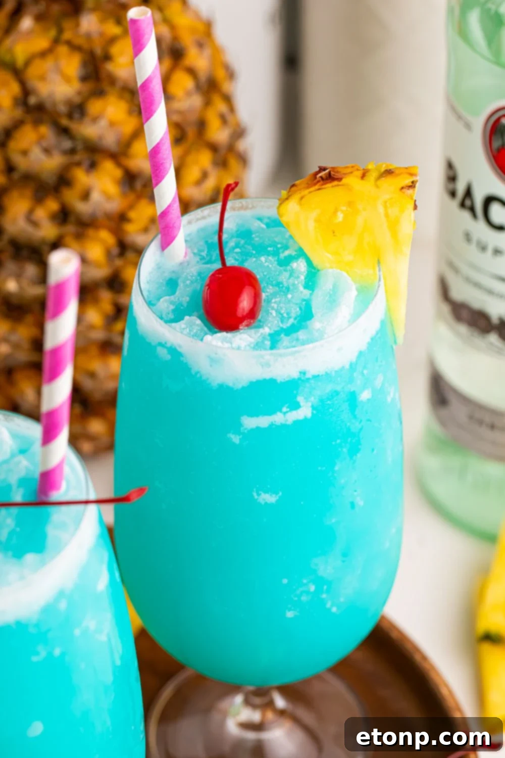 view of blue cocktails in glass with a pineapple on side and cherry in center of glass and pineapple and rum in background
