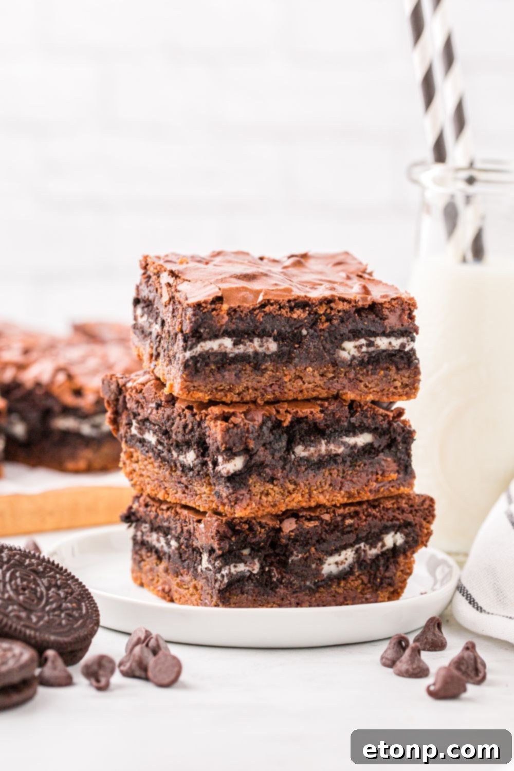 Slutty Brownies stacked on table with chocolate chips and Oreo cookies to the side and a glass of milk with straws in it 