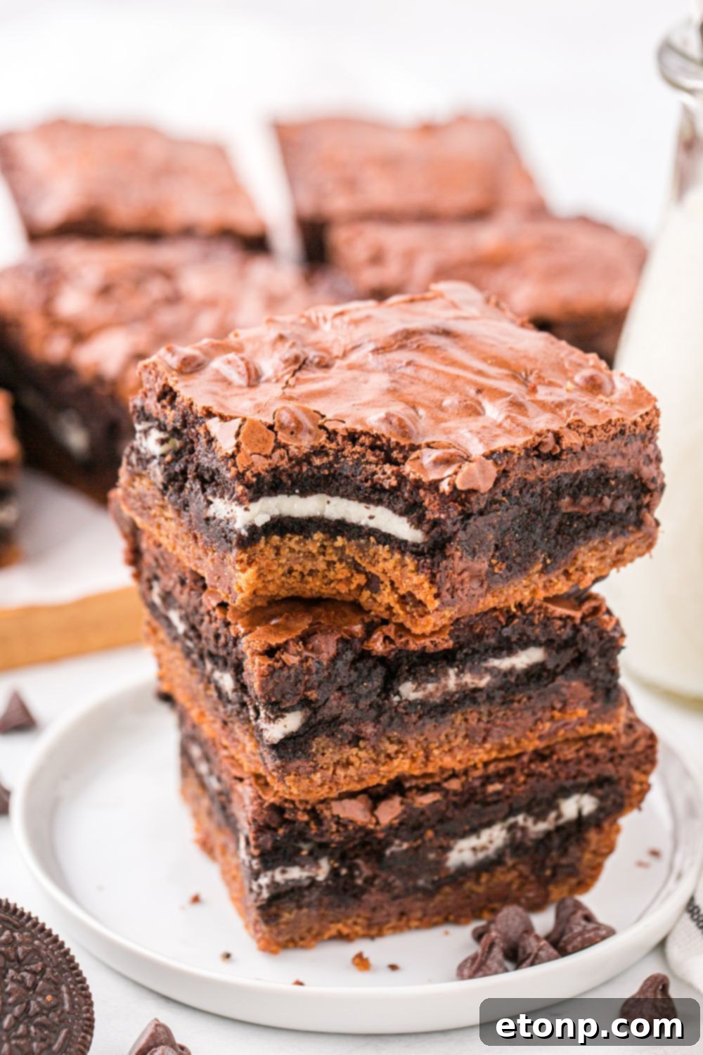 stack of Slutty Brownies on a white plate with a bite taken out of one, and behind a tray with the rest of the bars laid out 