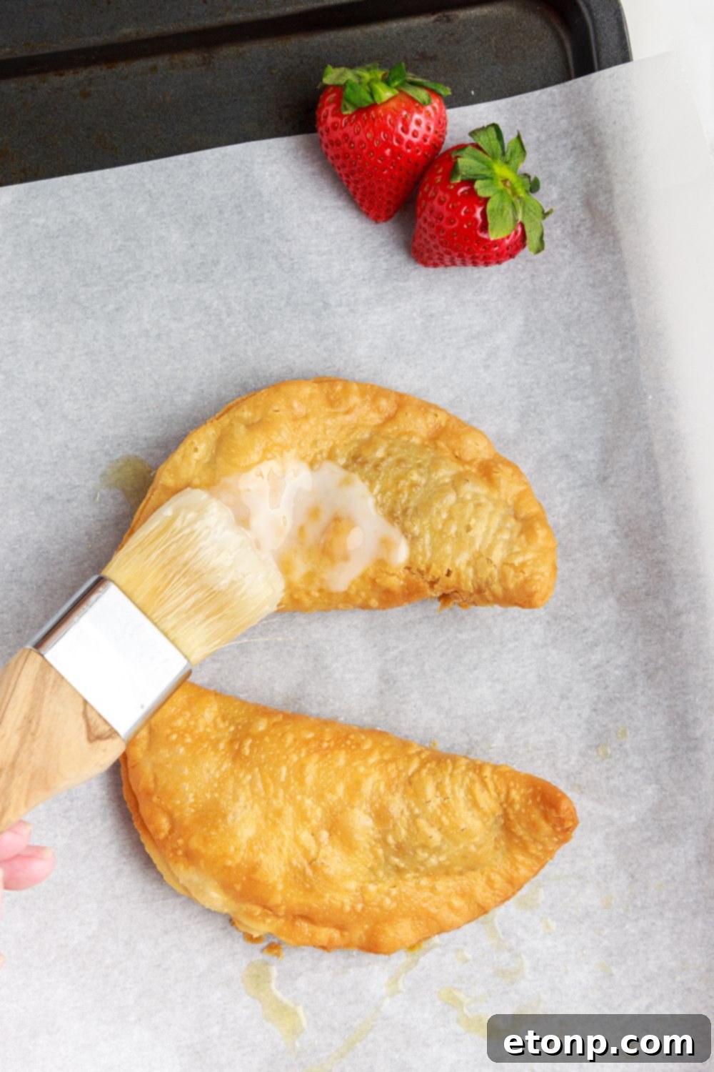 Deep Fried Strawberry Cheesecake Pies resting on a parchment-lined tray, being carefully brushed with a sweet glaze, enhancing their appearance.