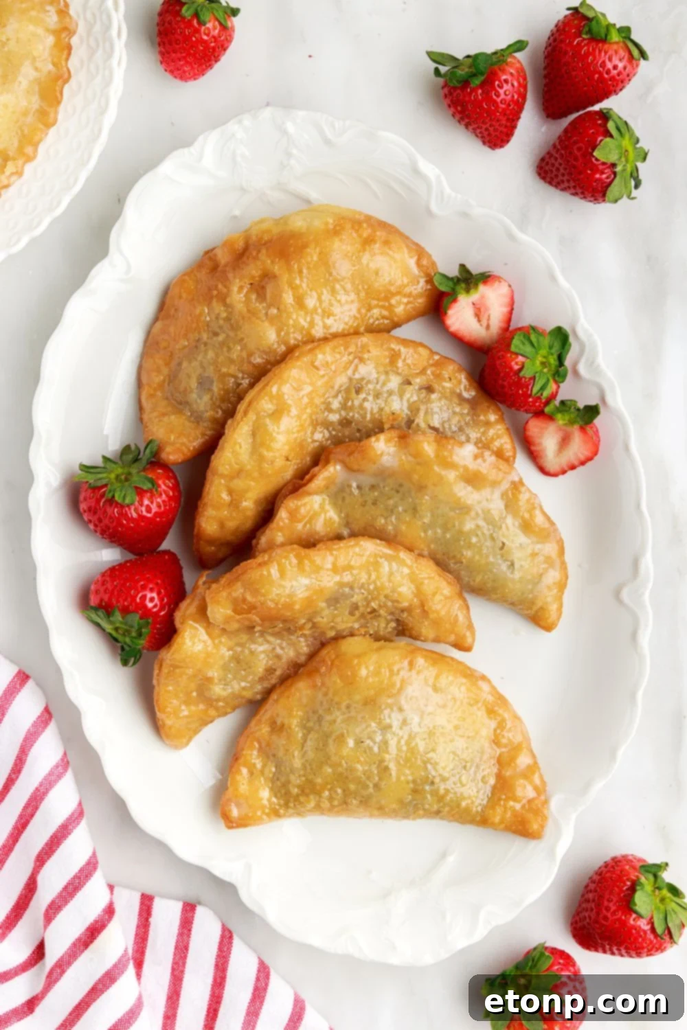 Deep Fried Strawberry Cheesecake Pies arranged attractively on a tray, with fresh strawberries scattered around the table and a red striped towel elegantly placed to the side.