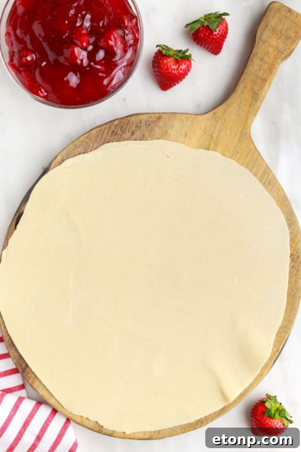 A pie crust unrolled and spread out on a cutting board, with a bowl of strawberry pie filling placed to the side, ready for assembly.