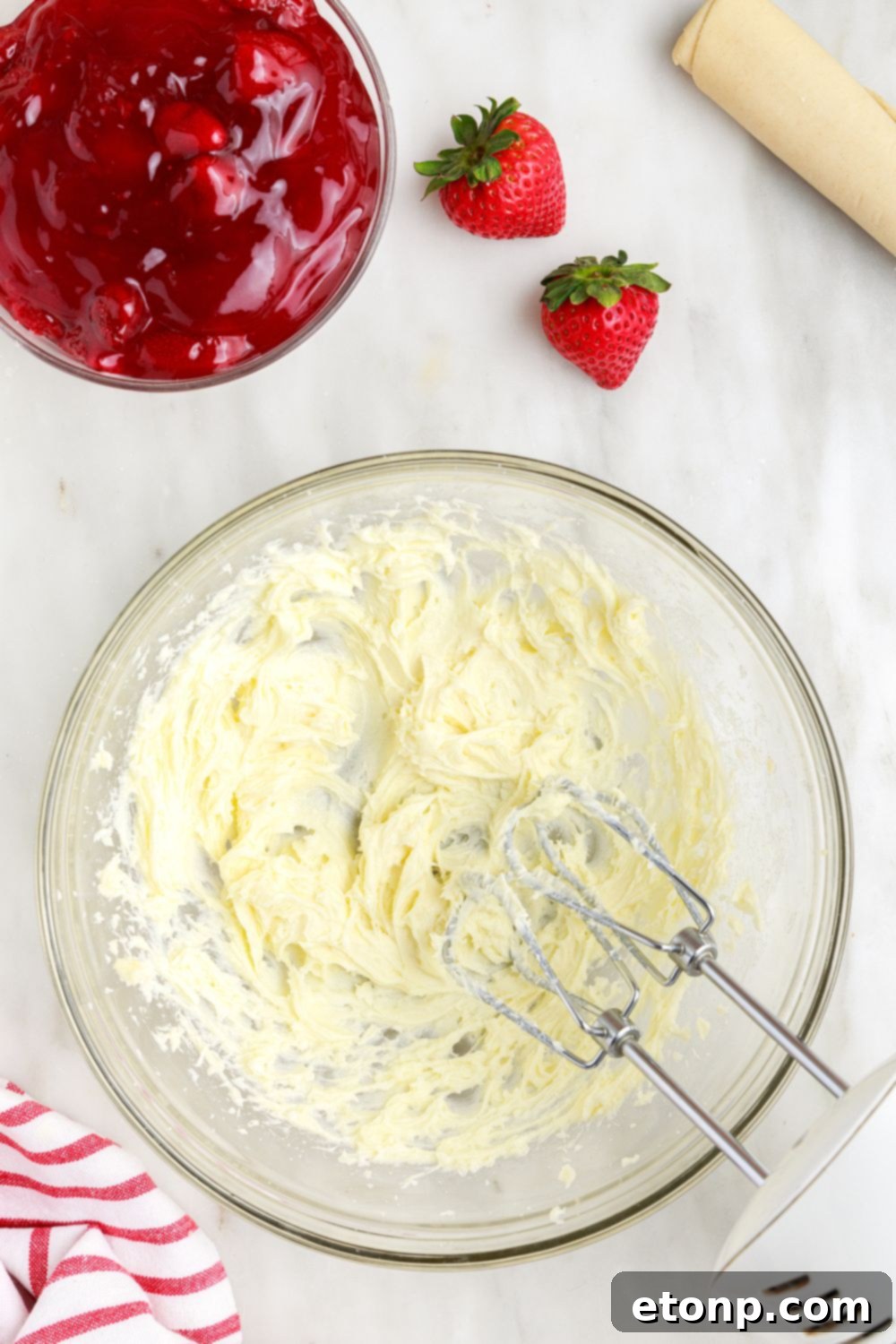 Cream cheese filling being mixed vigorously with a hand mixer in a bowl on a table, preparing for Deep Fried Strawberry Cheesecake Pies.