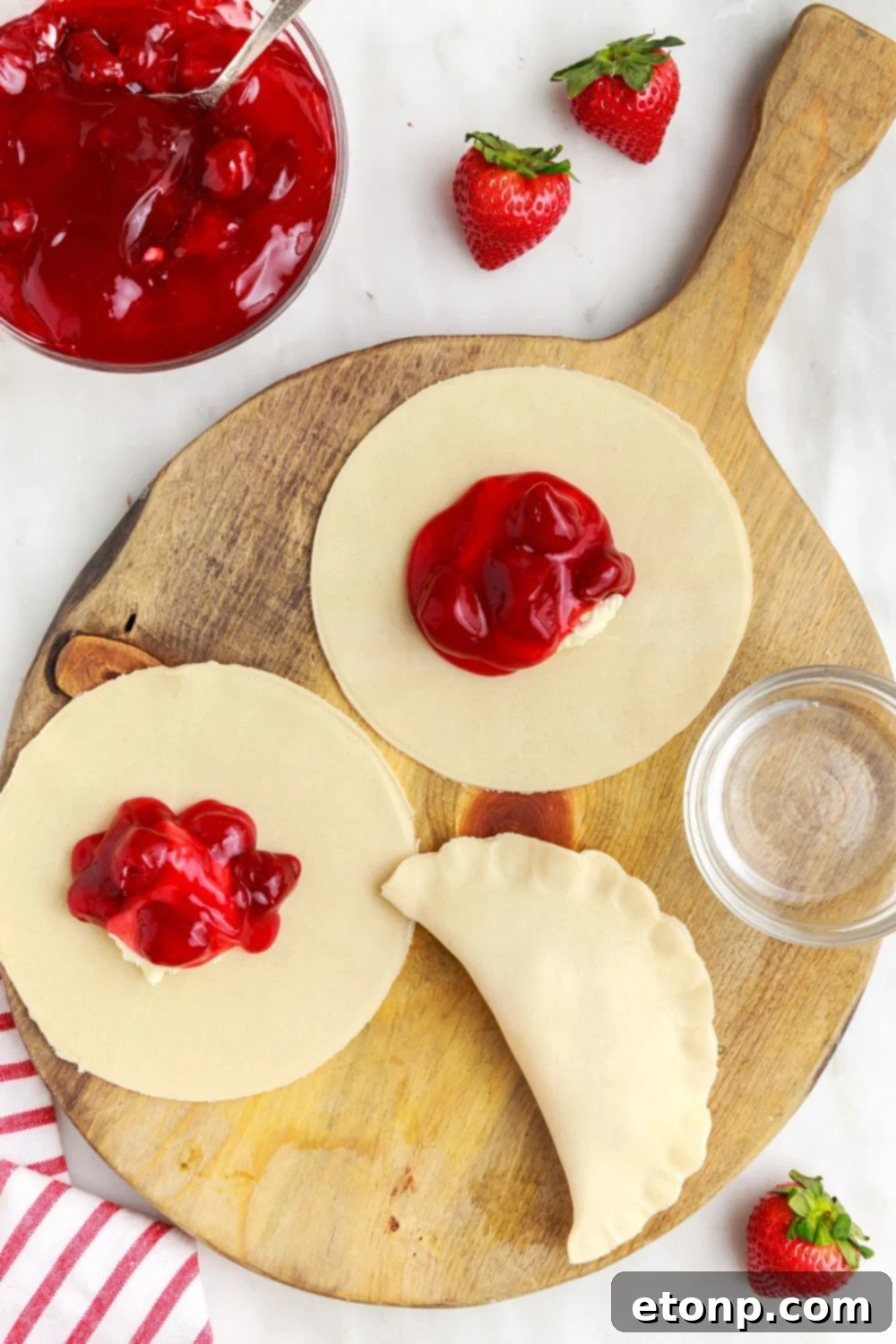The process of assembling strawberry hand pies, showing filling being spooned onto a pie crust laid out on a surface.