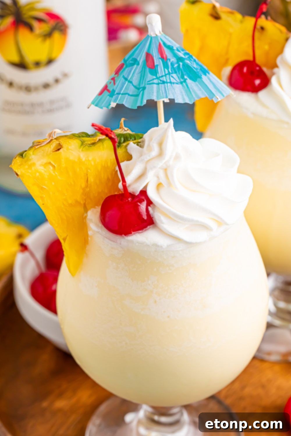 Frozen Malibu Piña Colada drinks on table with whipped cream, umbrella, and pineapple wedge in drink