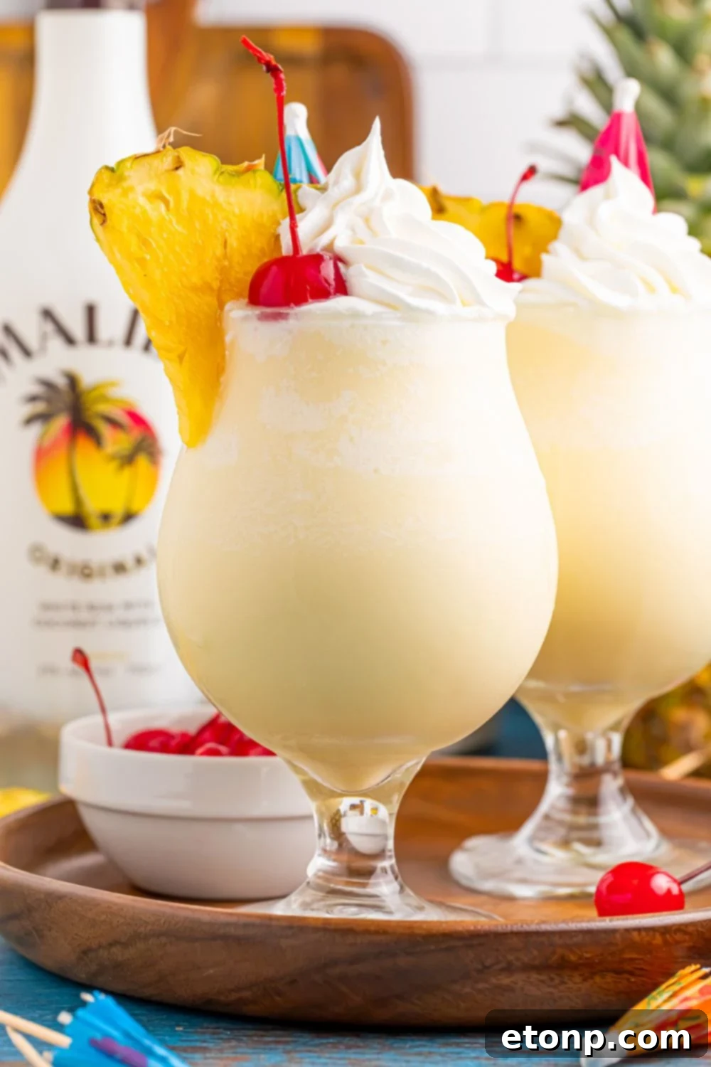 two Frozen Malibu Piña Coladas sitting on a tray with bowl of cherries, and a bottle of Malibu rum to the side