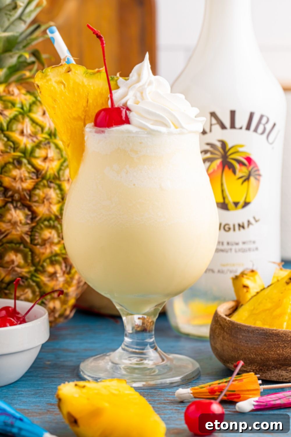 Frozen Malibu Piña Colada in a glass on table with pineapple slices and malibu and a whole pineapple to side on table