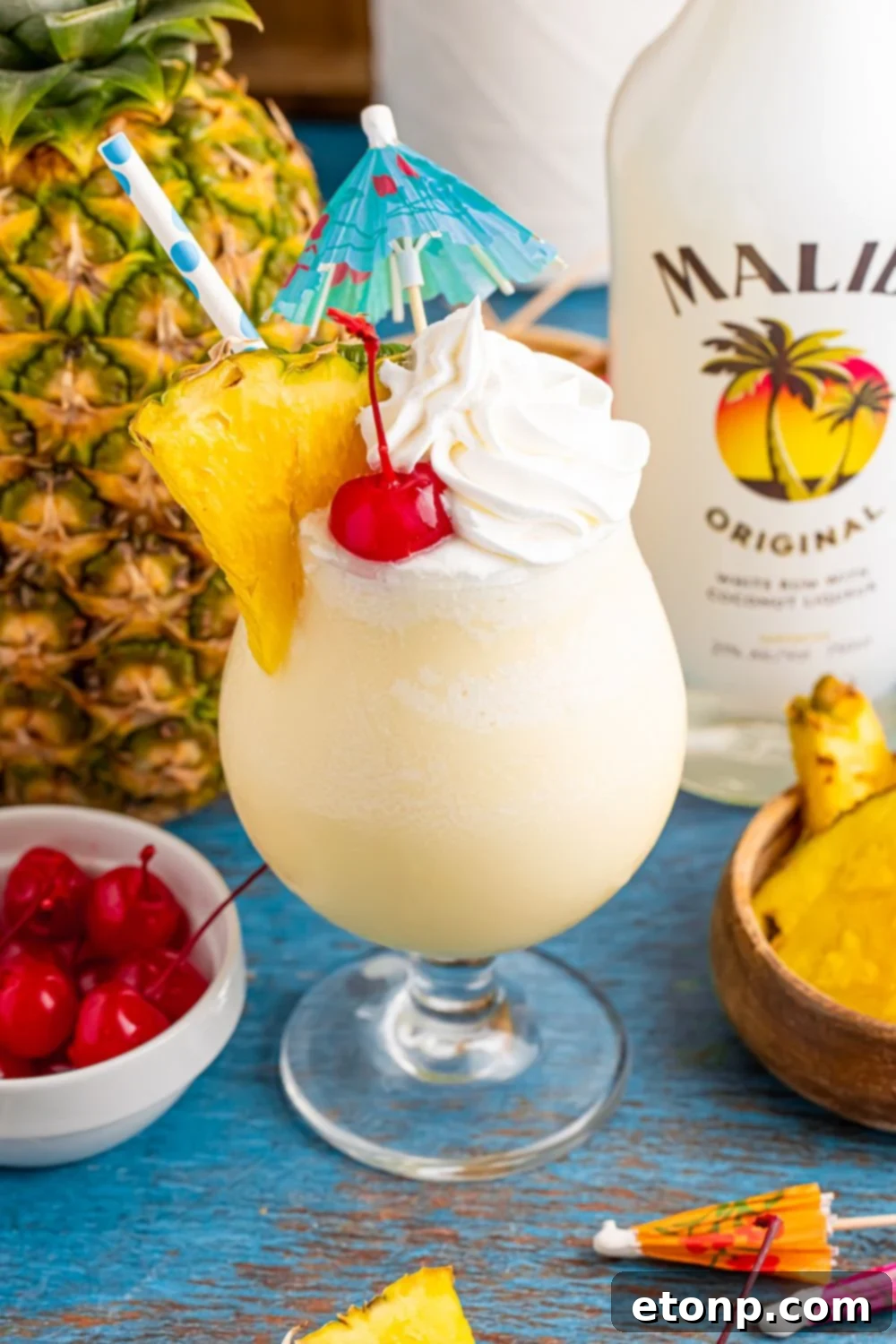 Frozen Malibu Piña Colada in a cocktail glass with a whole pineapple to side, bowl of cherries, and bottle of Malibu rum