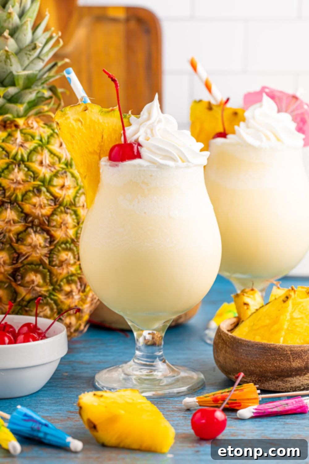 Frozen Malibu Piña Coladas on table with whole pineapple and cherries on table and umbrellas for drinks