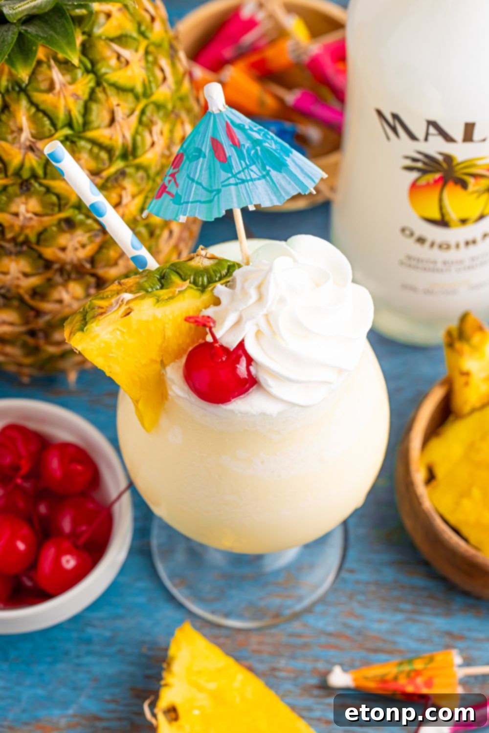 Frozen Malibu Piña Colada on table with a pineapple in the background and a bowl of cherries to side, and bottle of Malibu rum