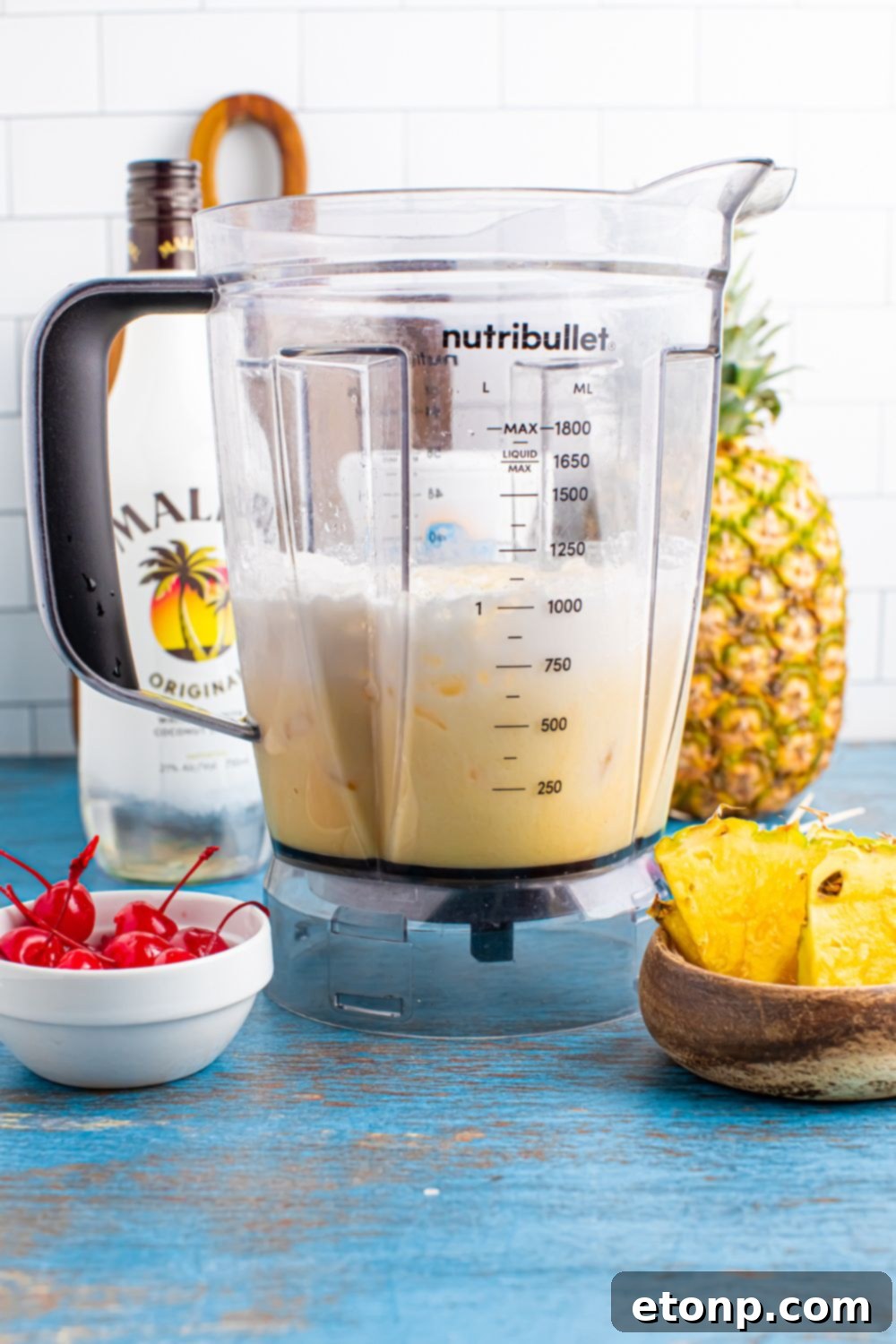 half mixed Frozen Malibu Piña Colada with a bowl of cherries and a bowl of pineapple, and Malibu rum in a bottle on table