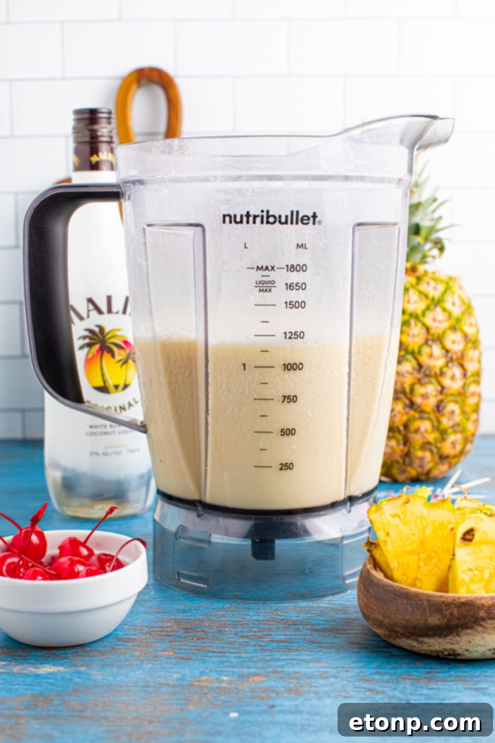 Frozen Malibu Piña Colada mixture in a blender sitting on table with pineapple, cherries, and pineapple