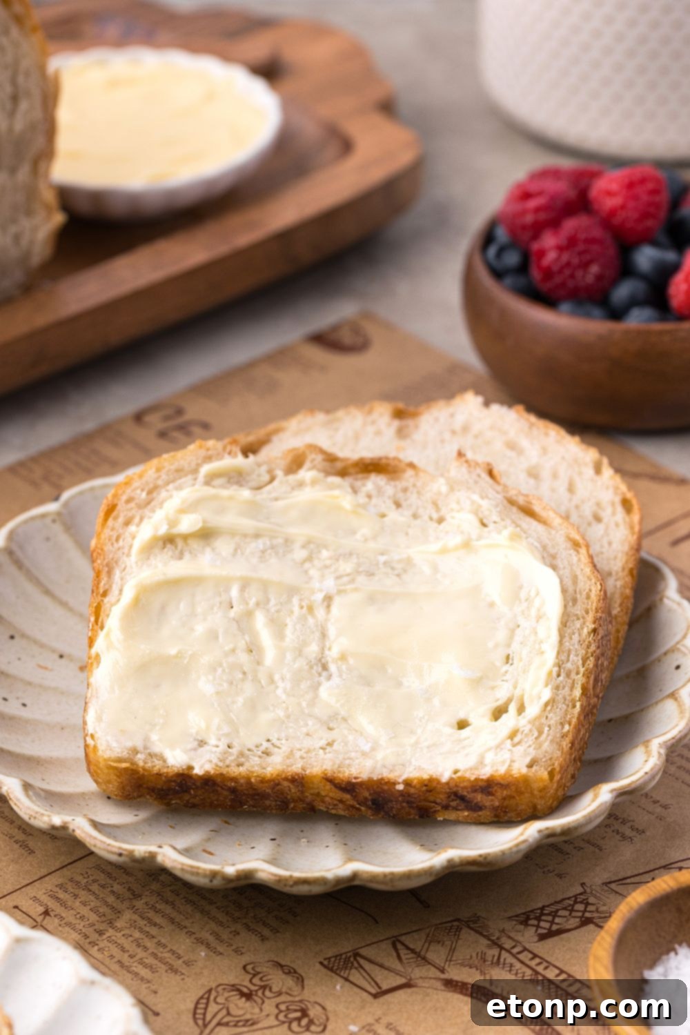 Homemade Sourdough Bread sliced and buttered sitting on plate with a bowl of fruit behind it on table 