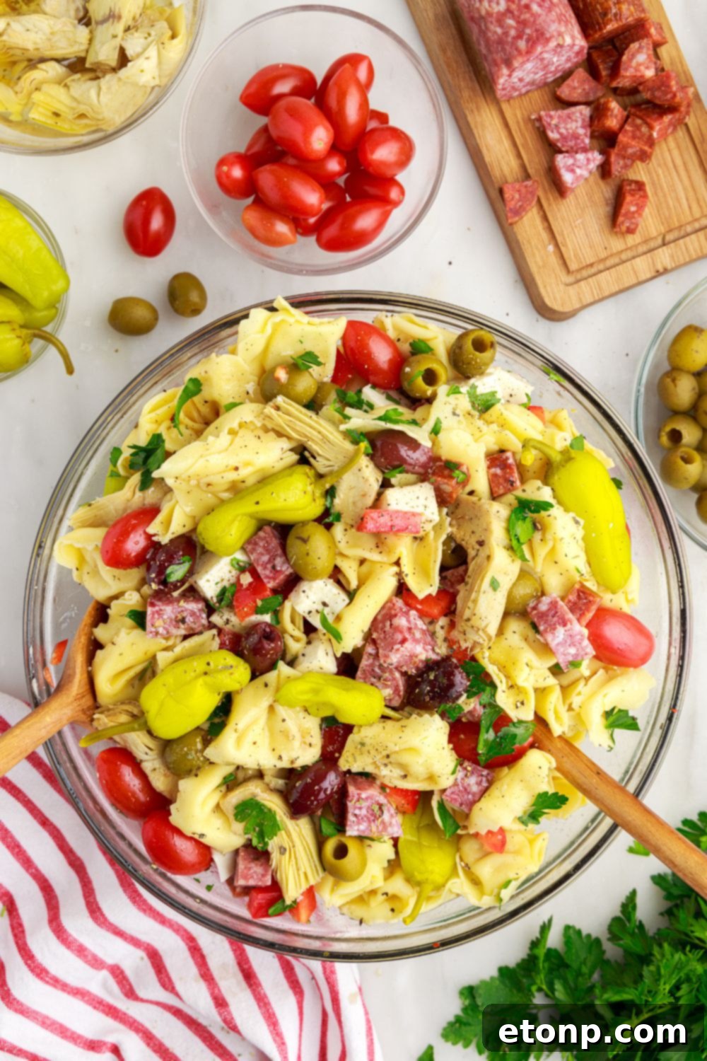 Antipasto Tortellini Pasta Salad on a table with ingredients scattered around and wooden serving spoons in the bowl.