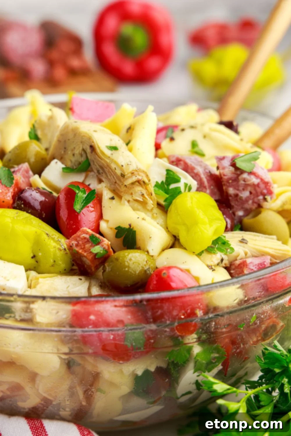 Side view of Antipasto Tortellini Pasta Salad in a glass bowl with serving spoons, showing the depth of the salad.