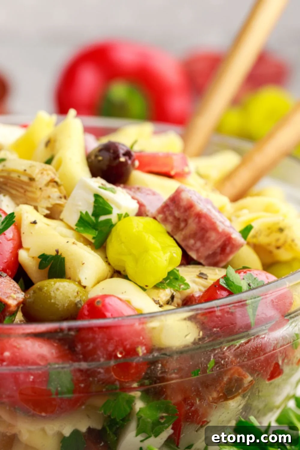Side view of Antipasto Tortellini Pasta Salad in a glass bowl with spoons and fresh vegetables around it.