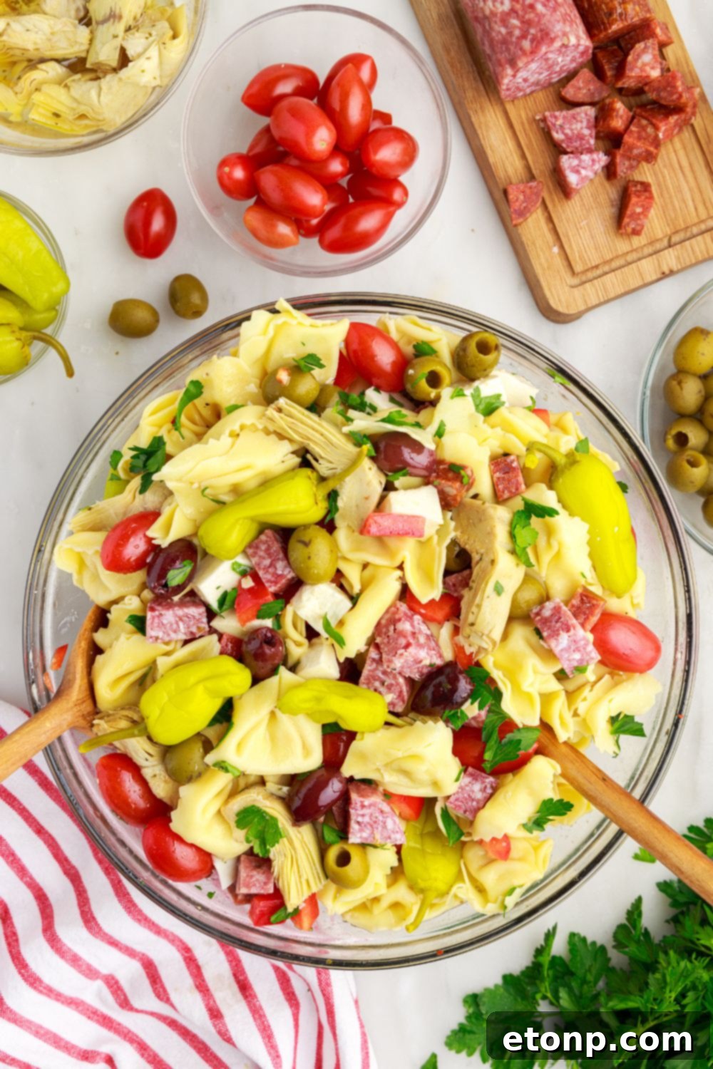 Antipasto Tortellini Pasta Salad in a glass bowl, with fresh bell peppers, tomatoes, and olives artfully arranged around it on a wooden table.