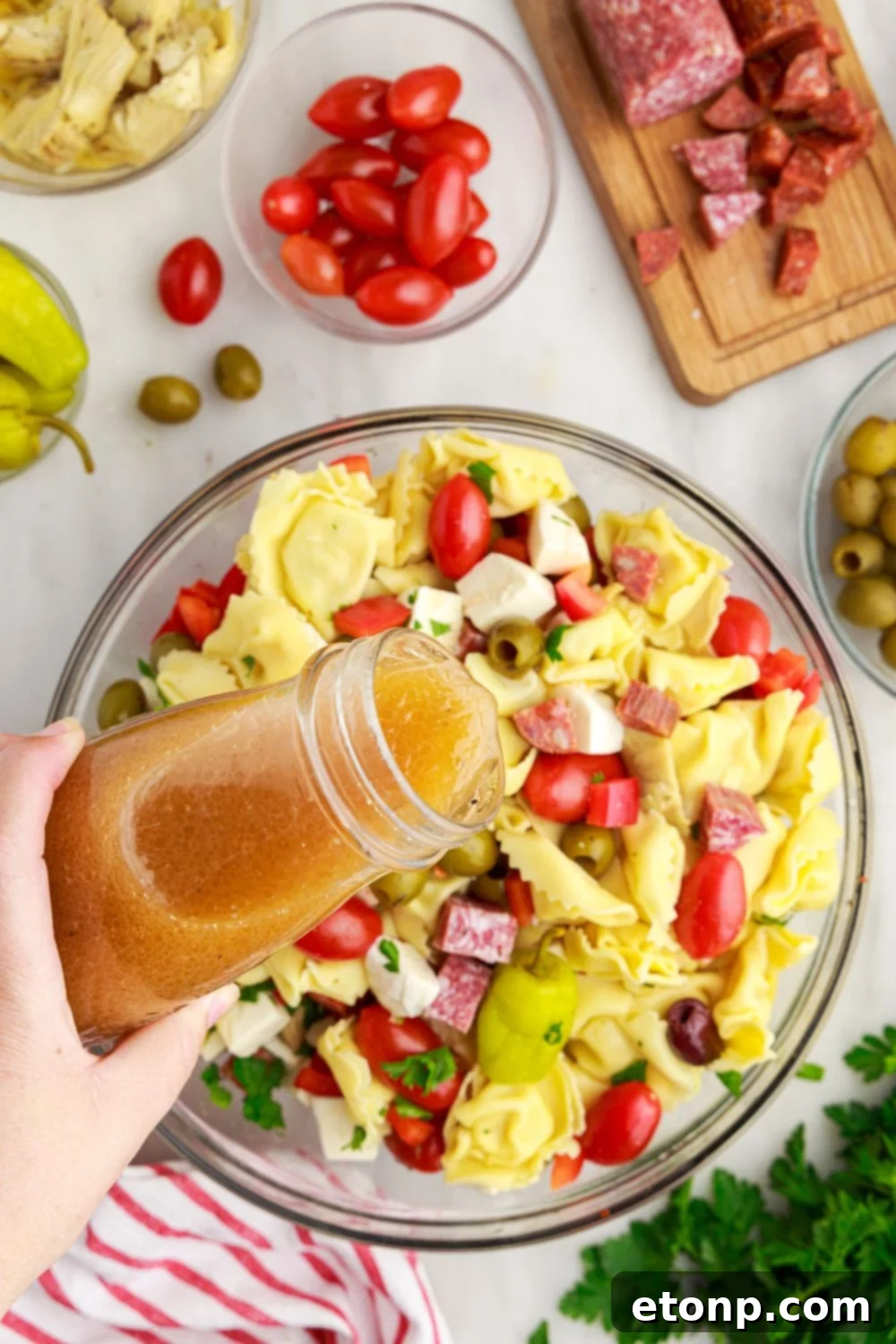 Zesty Italian dressing being poured over the Antipasto Tortellini Pasta Salad ingredients in a glass bowl.