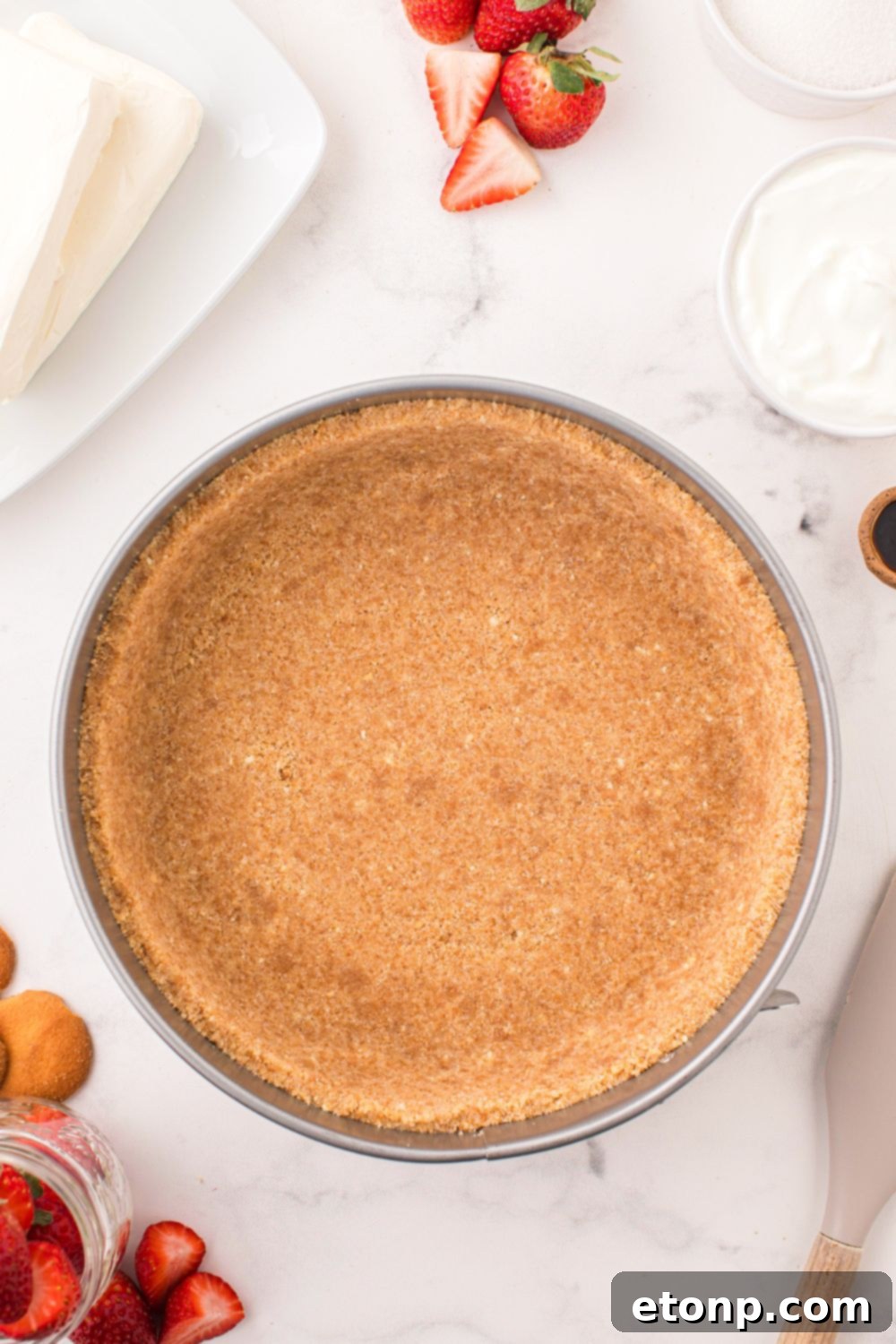 A perfectly pressed vanilla wafer crust resting in a pie plate, surrounded by fresh strawberries and other ingredients, awaiting the cheesecake filling.