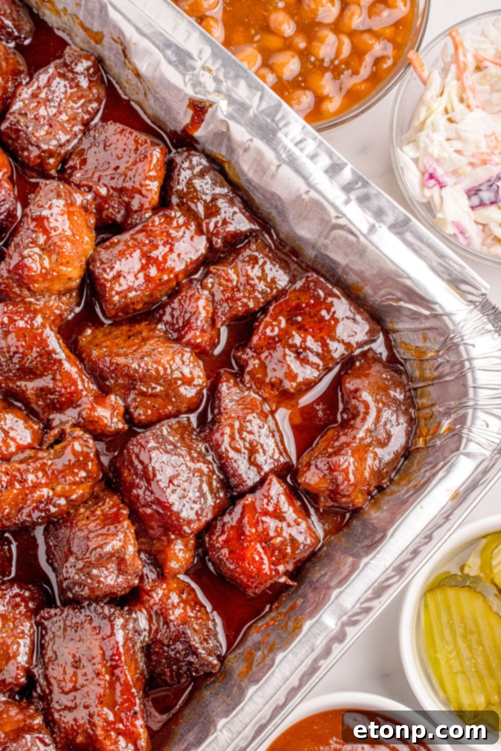 Chuck Roast Burnt Ends in a foil pan on table with beans and coleslaw in bowls to the side of the pan