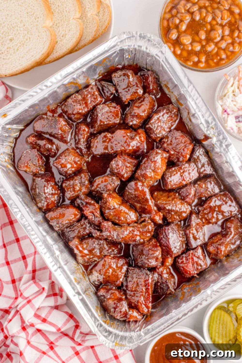Chuck Roast Burnt Ends in a foil baking pan on table with bread, beans, ketchup, and pickles in bowls around table